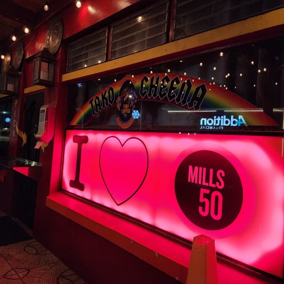 The exterior of Tako Cheena at night, with a neon pink "I ❤️ Mills 50" sign in the window.