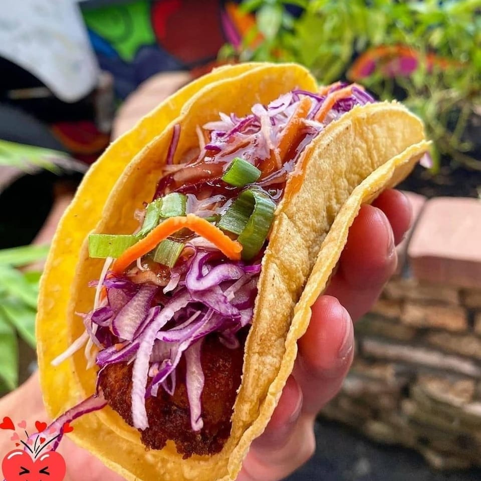 A hand holds a Tako Cheena Asian-fusion taco filled with crispy chicken, purple cabbage, and sauce.