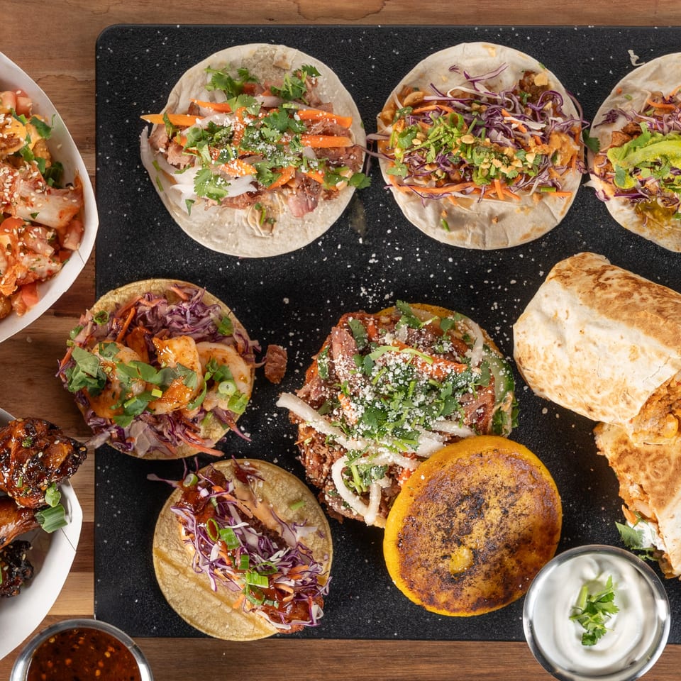 Overhead view of various Asian-fusion tacos, burritos, and sides from Tako Cheena on a dark surface.